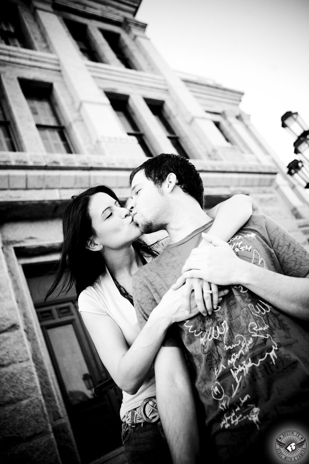 dark haired girl with nice facial bone structure wearing a low cut light short sleeve blouse and large belt embraces from behind while kissing a brunette guy wearing a fashionable grey t-shirt with an abstract white line design in front of the East side of the Texas Capital Building in this adventurous engagement session in Austin.